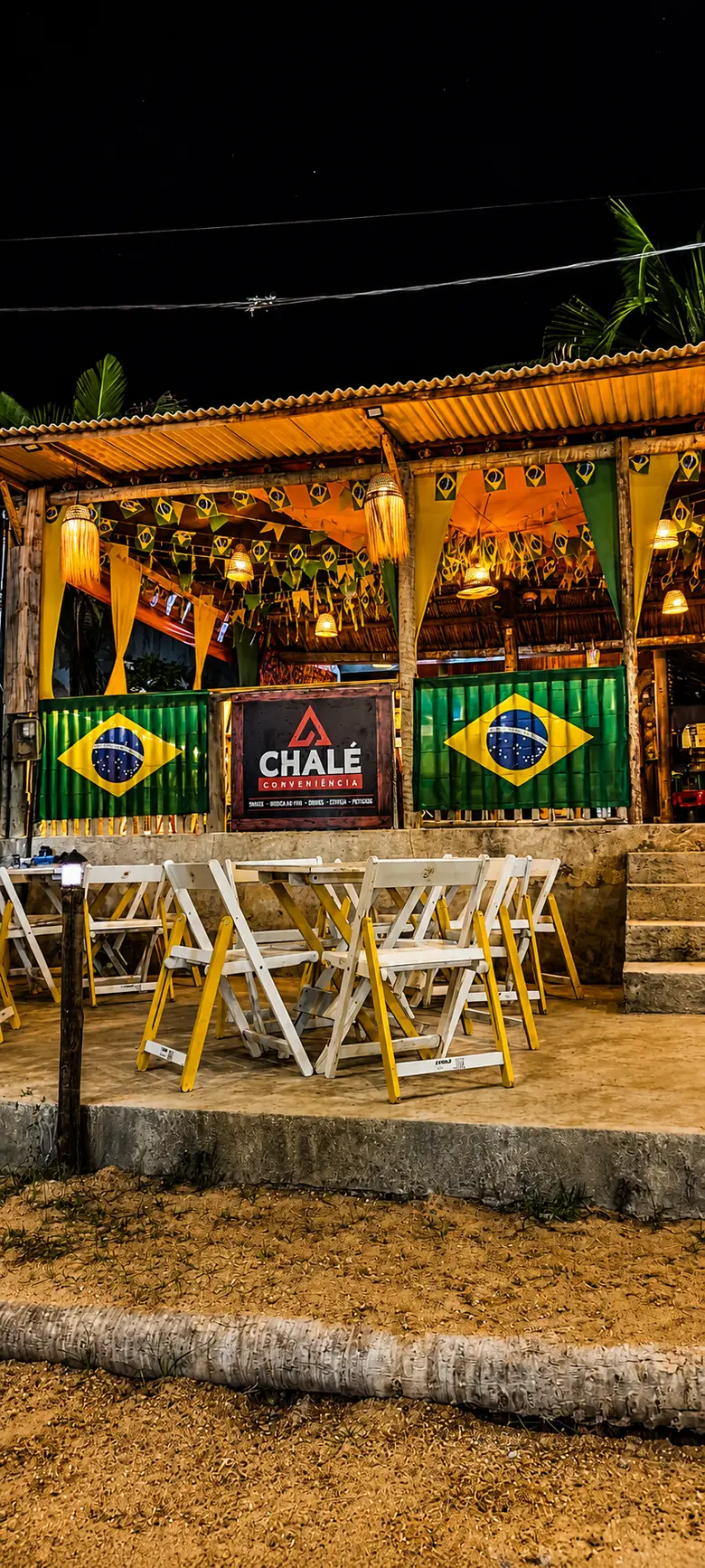 Fachada do Chalé Conveniência à noite, com o letreiro aceso e as cadeiras na areia